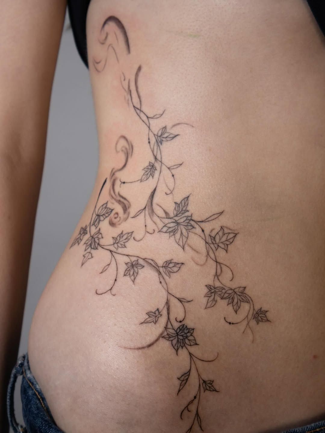 A delicate, fine-line tattoo of trailing ivy vines and small leaves winding along the side of a person's torso.