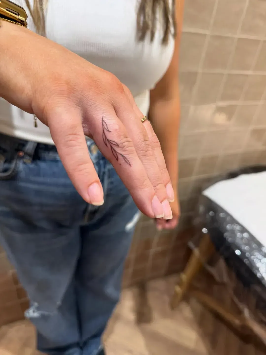 A hand shows a delicate, fine-line leaf tattoo on the side of the ring finger.