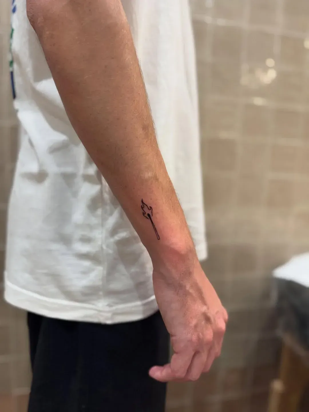 A small, minimalist black ink tattoo of a burning matchstick on the underside of a person's forearm.