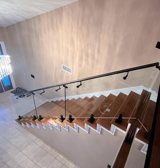 Staircase with wooden steps, glass panels, black handrail. Beige walls. Bright chandelier in the background.