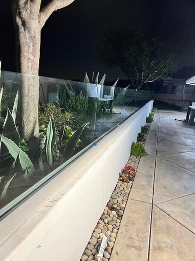 Outdoor view at night: white wall with glass railing next to a garden bed with rocks, plants, and a tree.