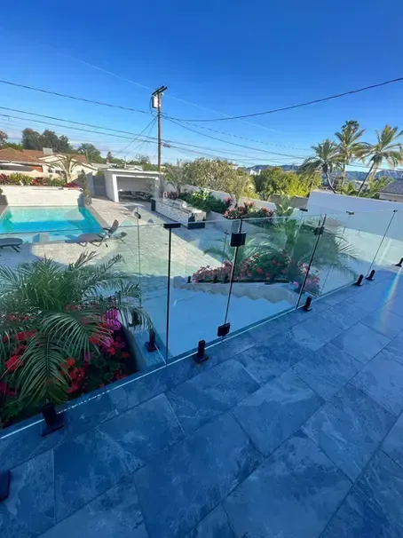 Pool area with glass fencing. Blue tiled patio, pool, and landscaping. Clear sky.