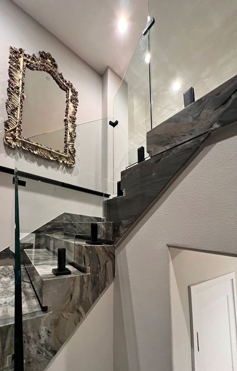 Staircase with gray stone steps, glass railing, and ornate gold-framed mirror on a white wall.