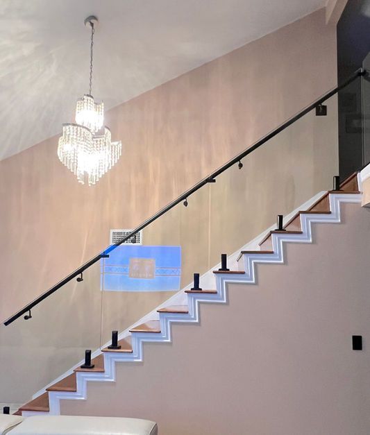 Staircase with glass railing, black handrail, zigzag-shaped white risers, and a chandelier in a light pink room.