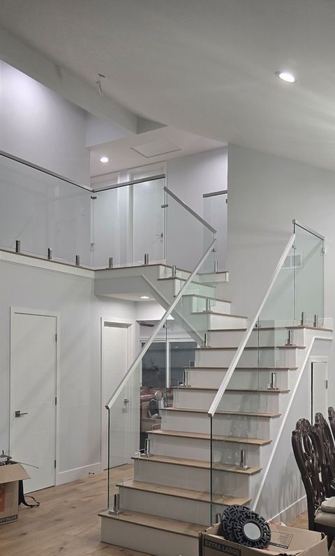 Staircase with glass railing and white risers. Natural wood stairs, white walls, and a light wooden floor.
