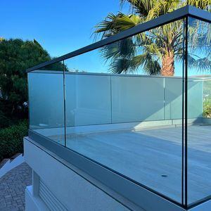 Modern glass balcony with gray frame, overlooking trees under a blue sky.