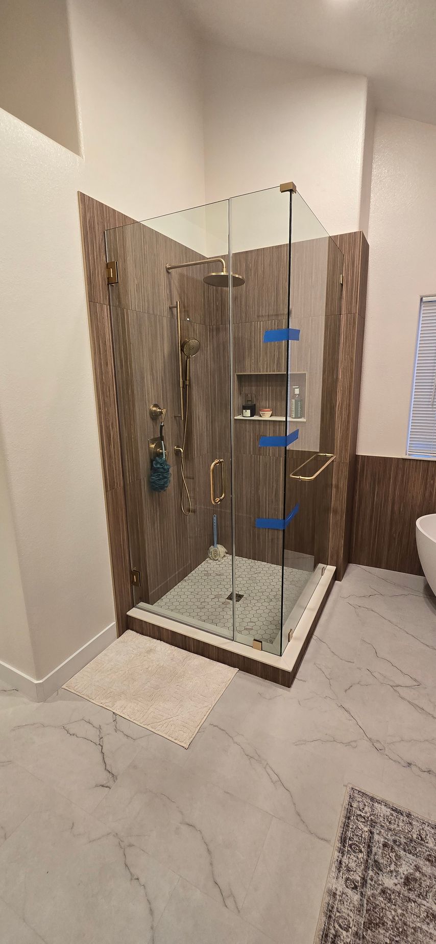 Bathroom with a glass shower enclosure featuring brown tiled walls, gold fixtures, and gray and white marbled flooring.