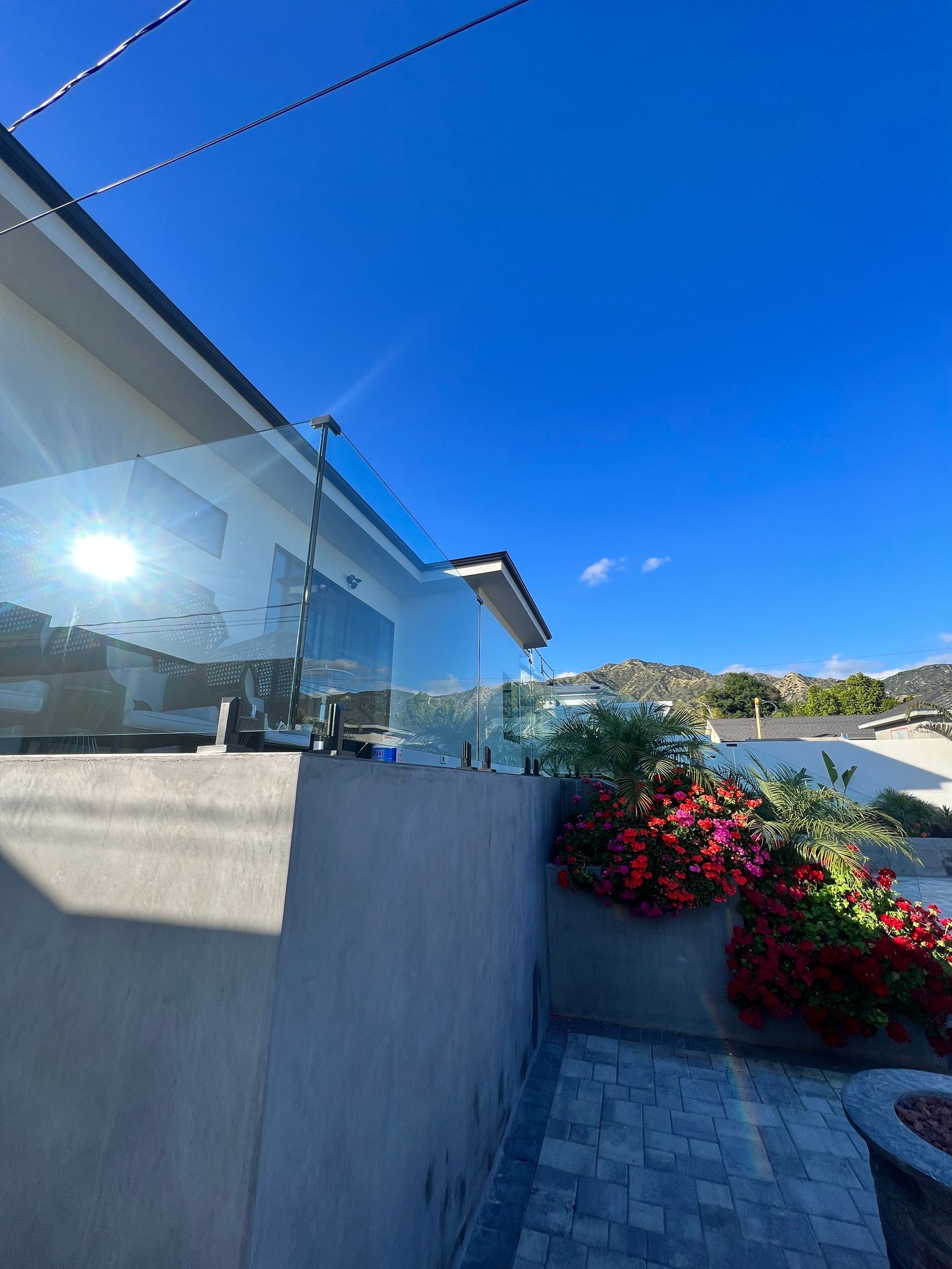 Glass railing on a cement wall next to red flowers under a bright blue sky.