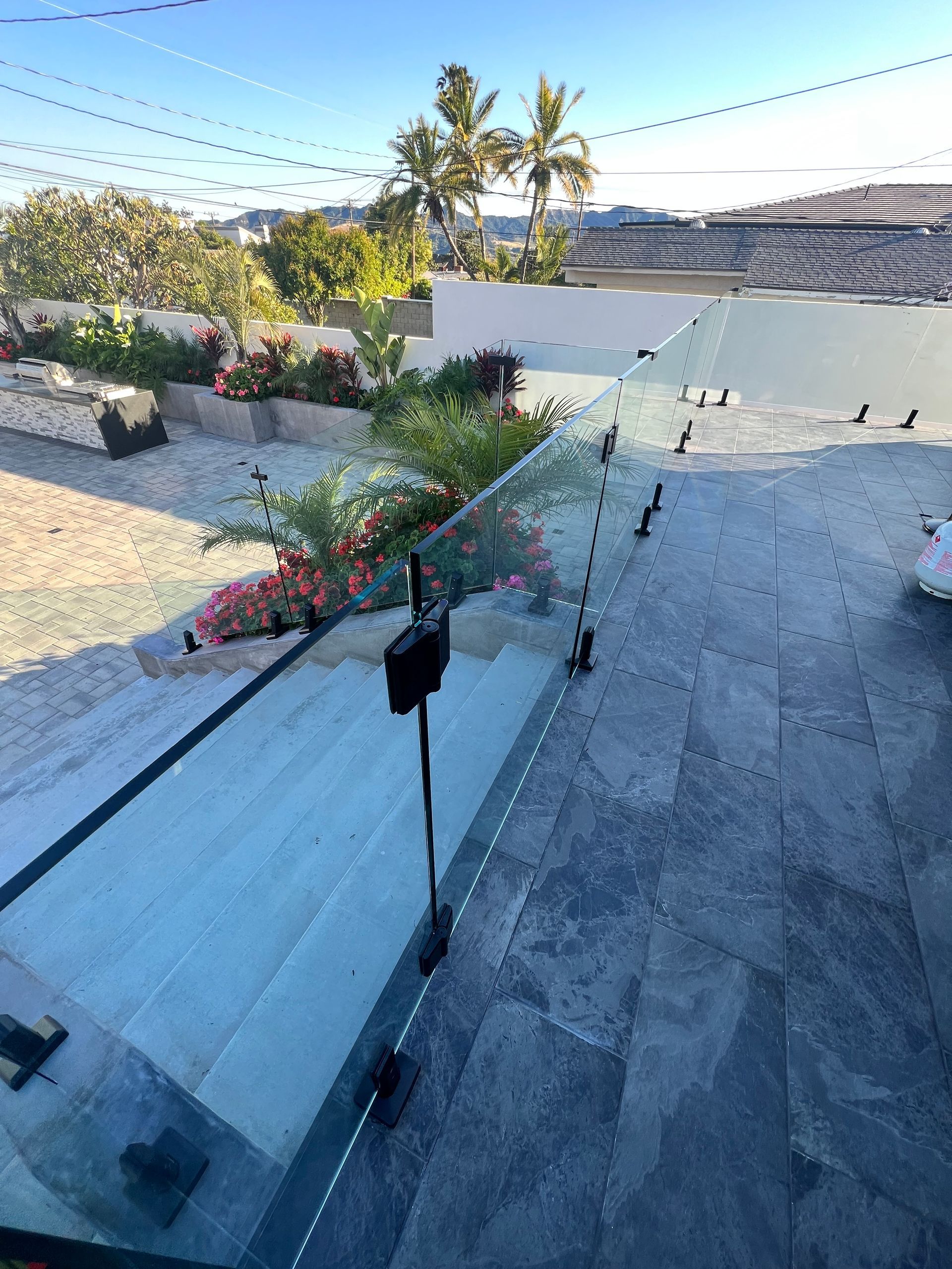 Glass railing on stone steps leading to a garden with pink flowers and palm trees under a bright sky.