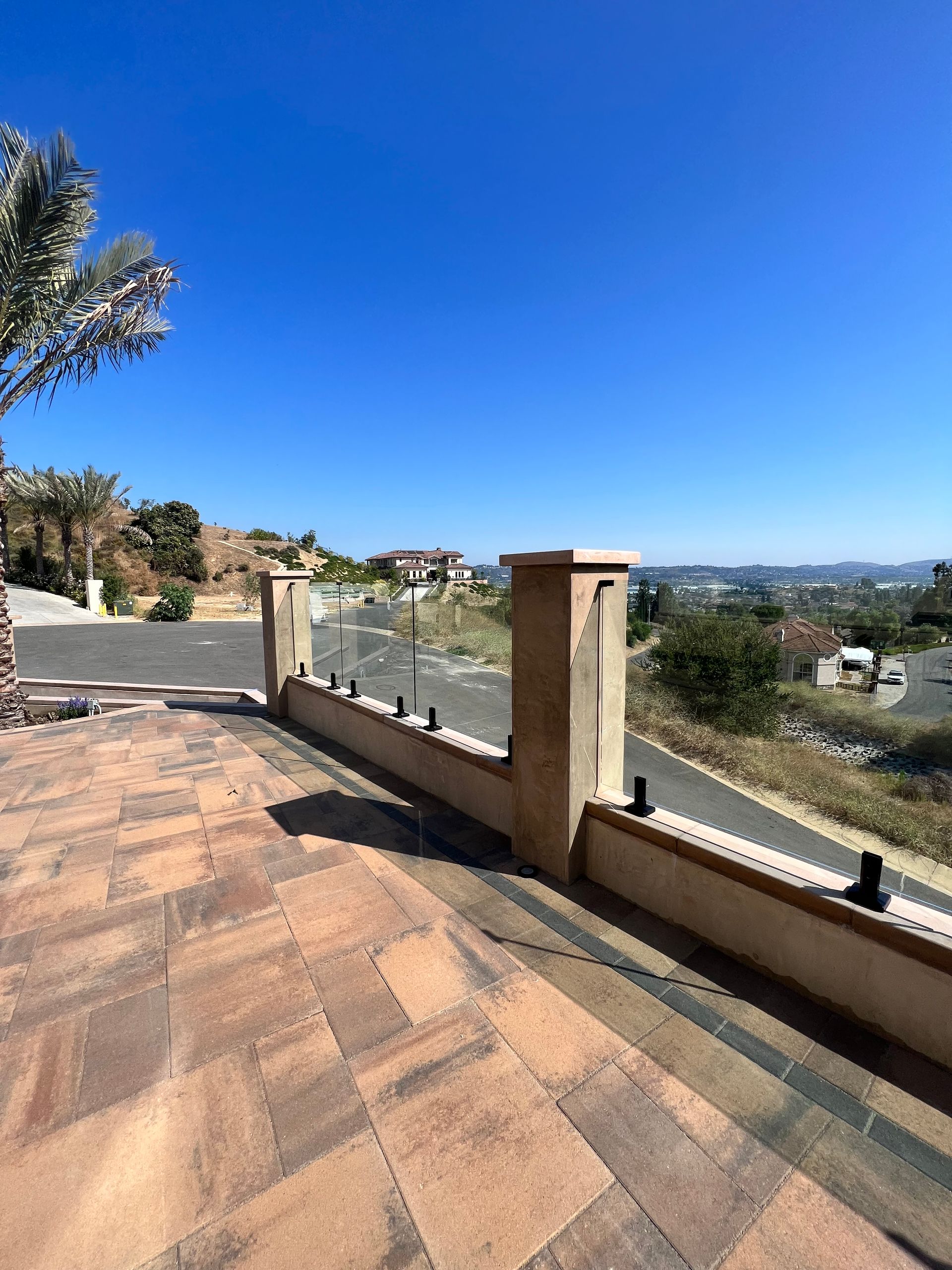 Patio with view: brown tile, cement barrier with glass, overlooking a road and houses, clear blue sky.