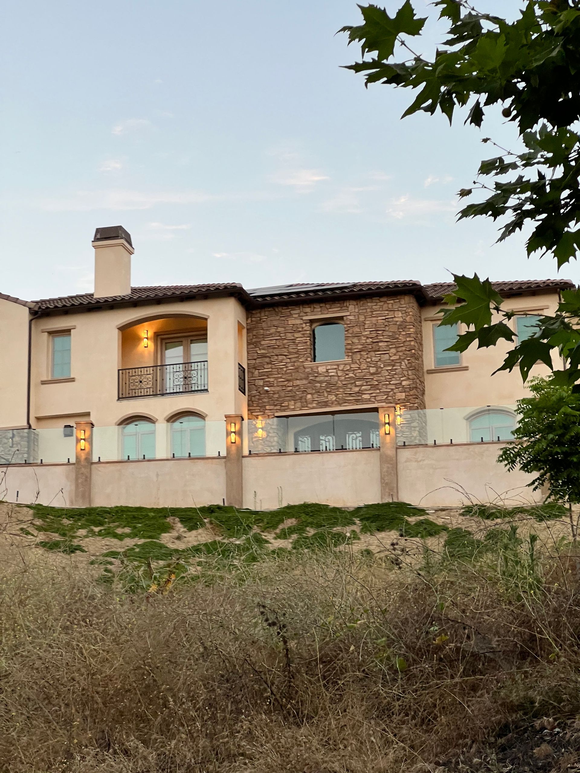Beige two-story house with stone facade, balcony, and glass railing on a hillside; dry grass in foreground.