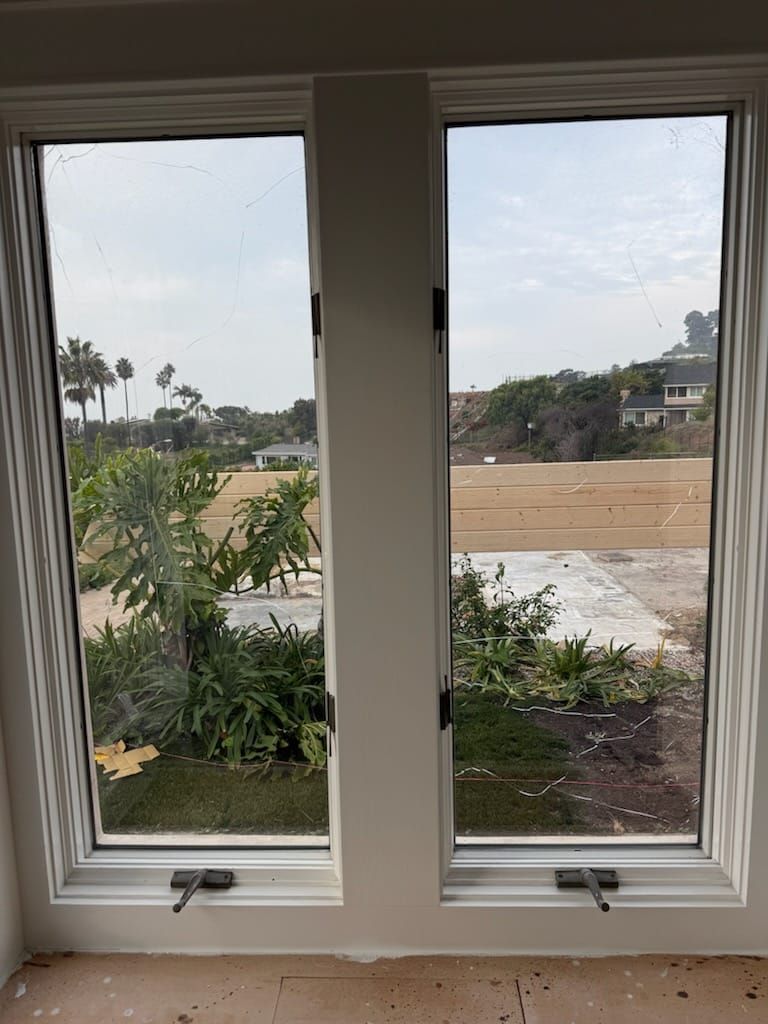 Two white-framed windows with views of greenery and an outdoor space under an overcast sky.