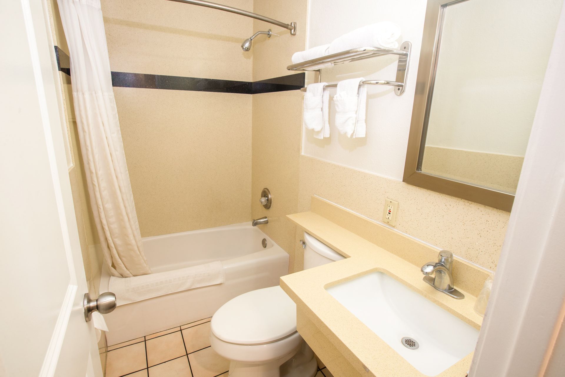 A small hotel bathroom with a bathtub, toilet, sink, and mirror. White and beige tones.