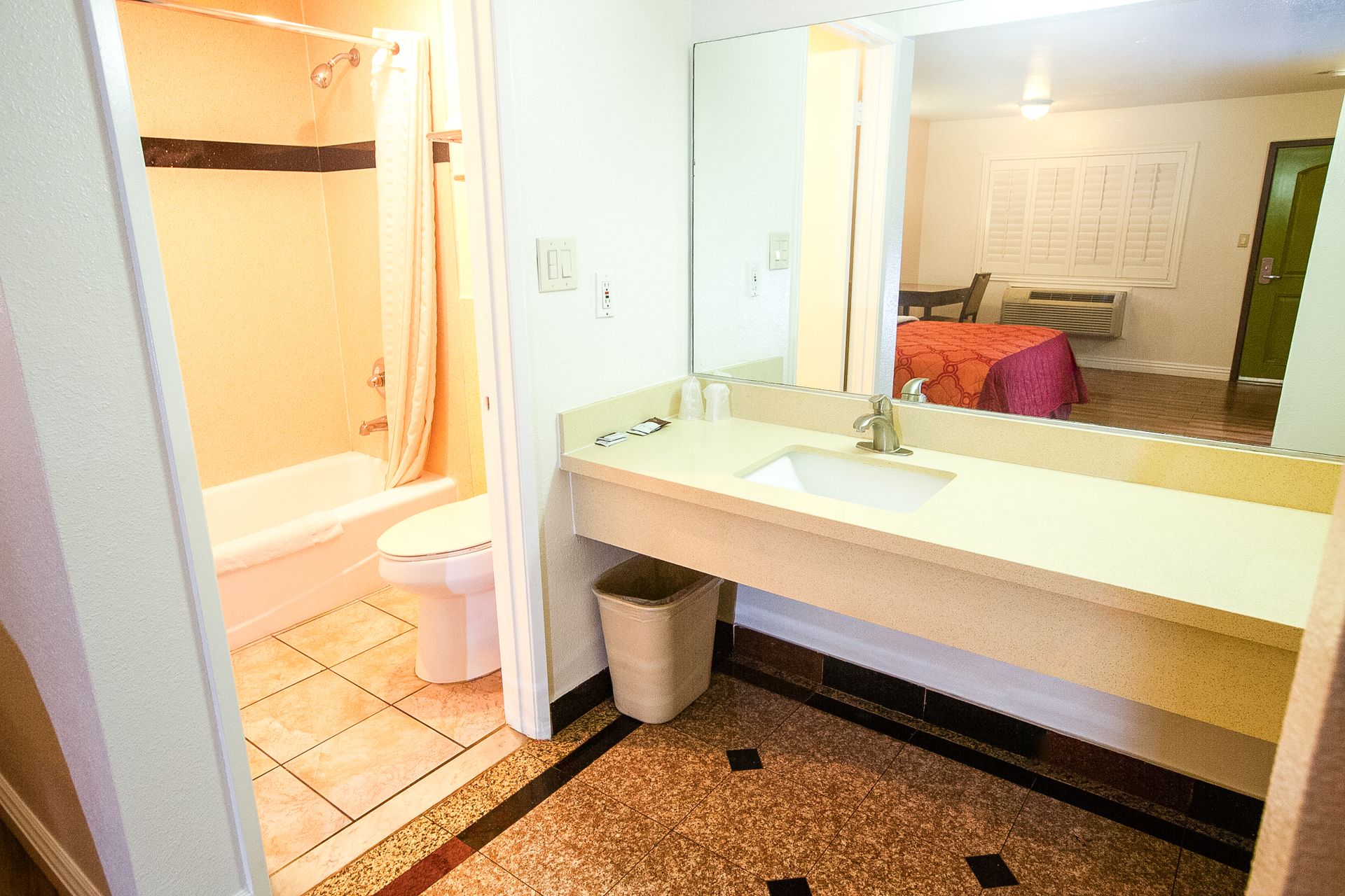 Bathroom with beige tub/shower, toilet, sink, and mirror. View into bedroom with bed and desk.