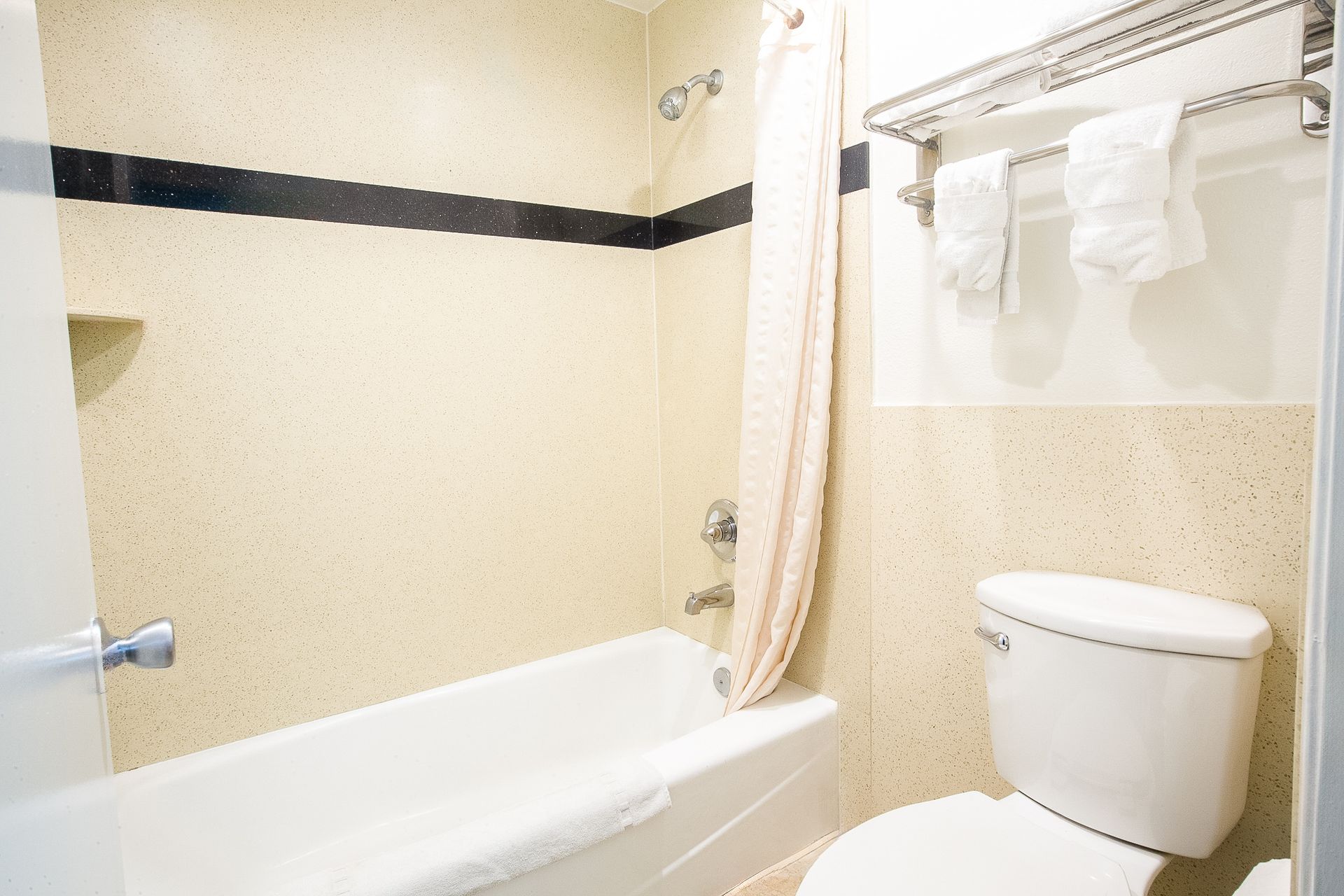 Bathroom with white tub, toilet, and towels on a rack. Beige walls, shower curtain, and black stripe.