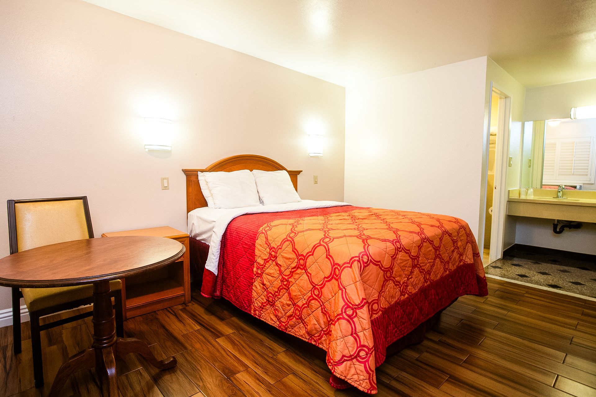 Hotel room with a bed, table, chair, and bathroom visible in the background. Red and orange bedding, wooden floor.