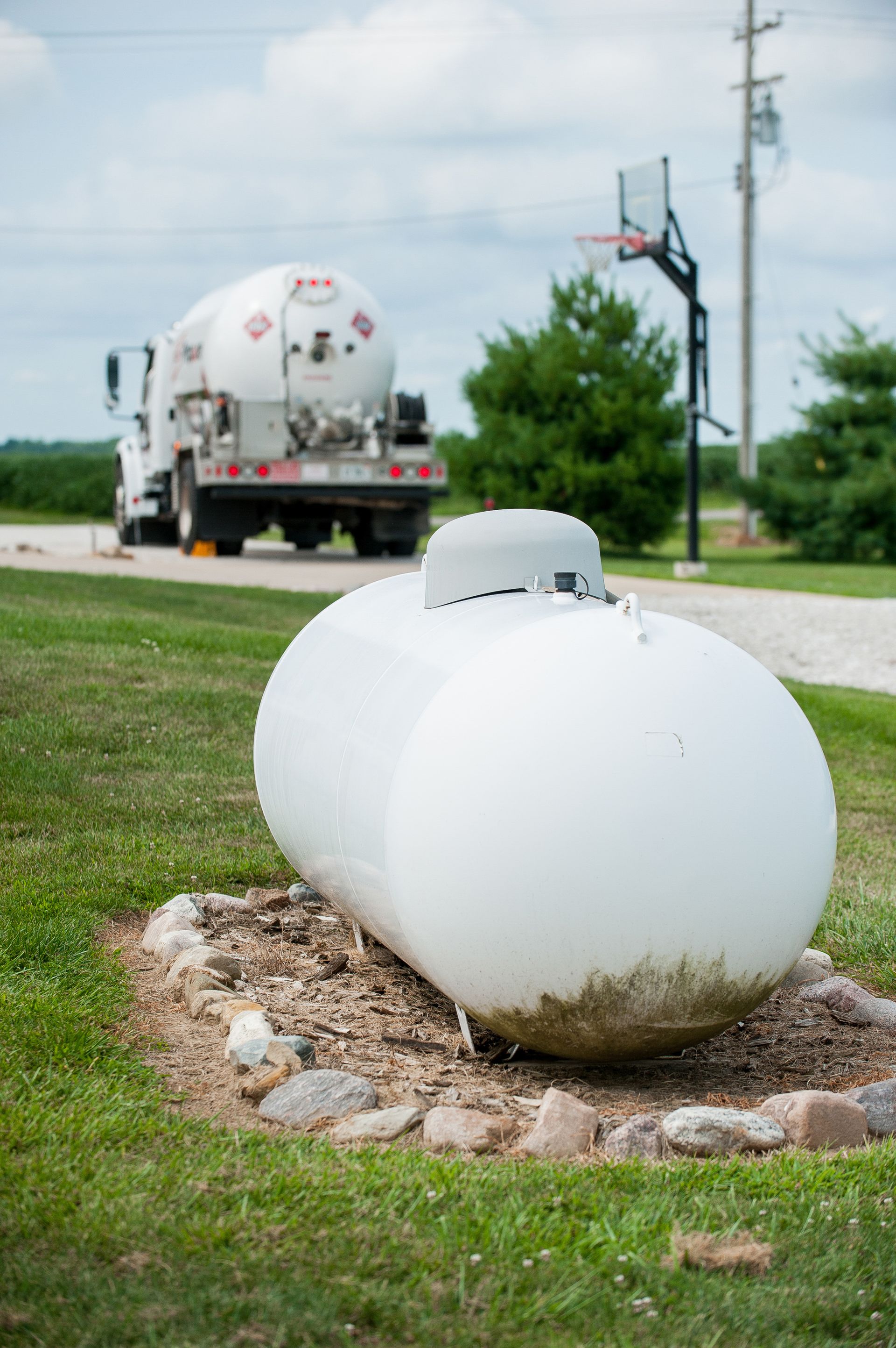 White propane tank on a gravel patch beside a road, with a fuel truck in the background.