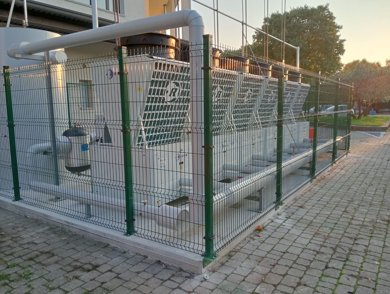 Attrezzature industriali all'aperto, recintate con staccionata verde, con tubi e cavi in un'area pavimentata al tramonto.
