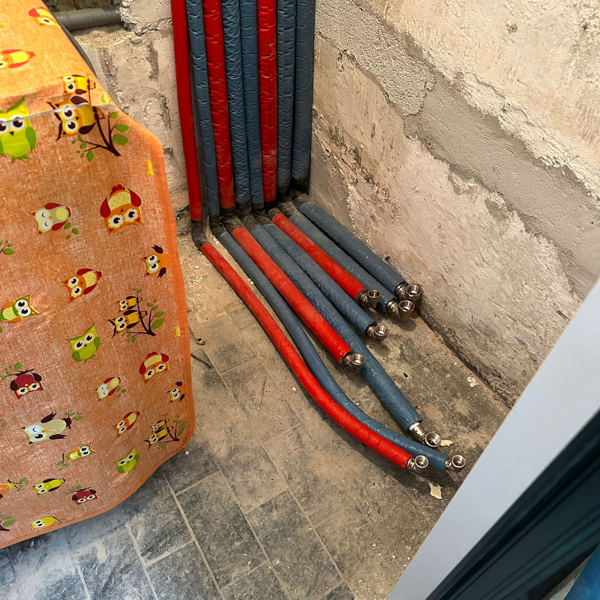 Tubi rossi e blu emergono da un muro. Il pavimento è piastrellato di grigio. Un oggetto arancione decorato è inquadrato.