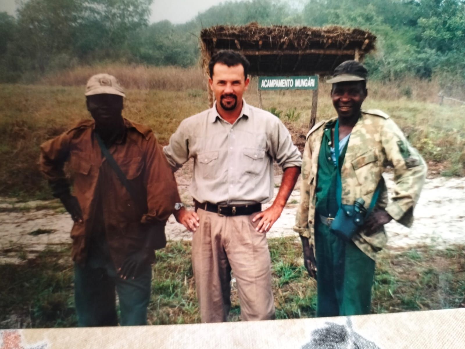Three men standing outdoors in a field, with a sign and stacked materials in the background.