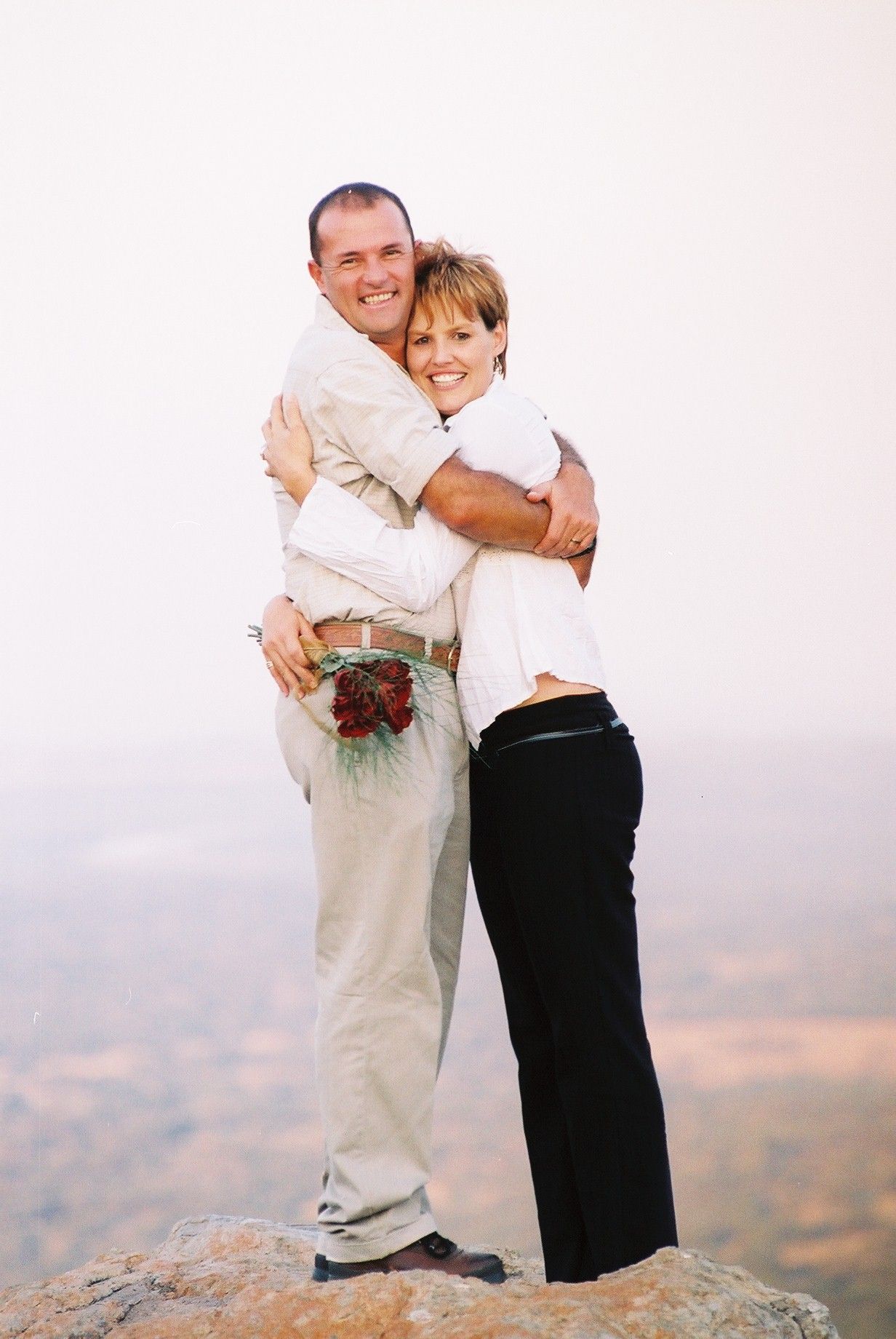 Two people embracing and smiling on a foggy hilltop, with a bouquet behind them.