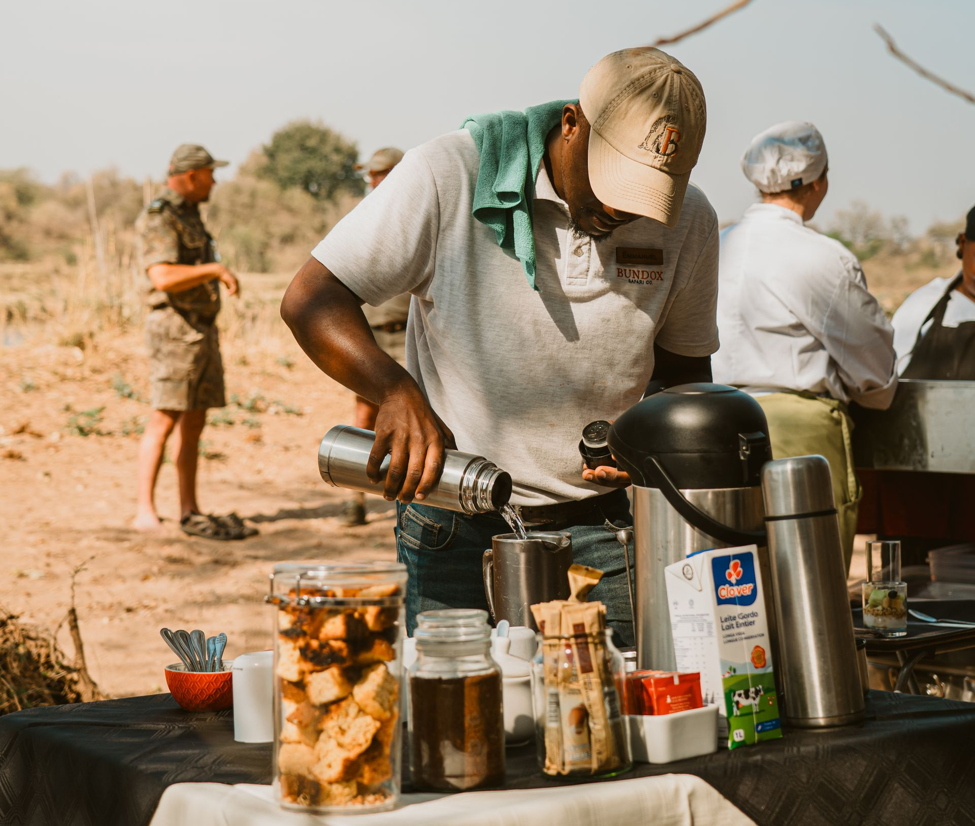 Person preparing drinks at an outdoor table in a dry, sunny setting, with jars and a coffee pot nearby