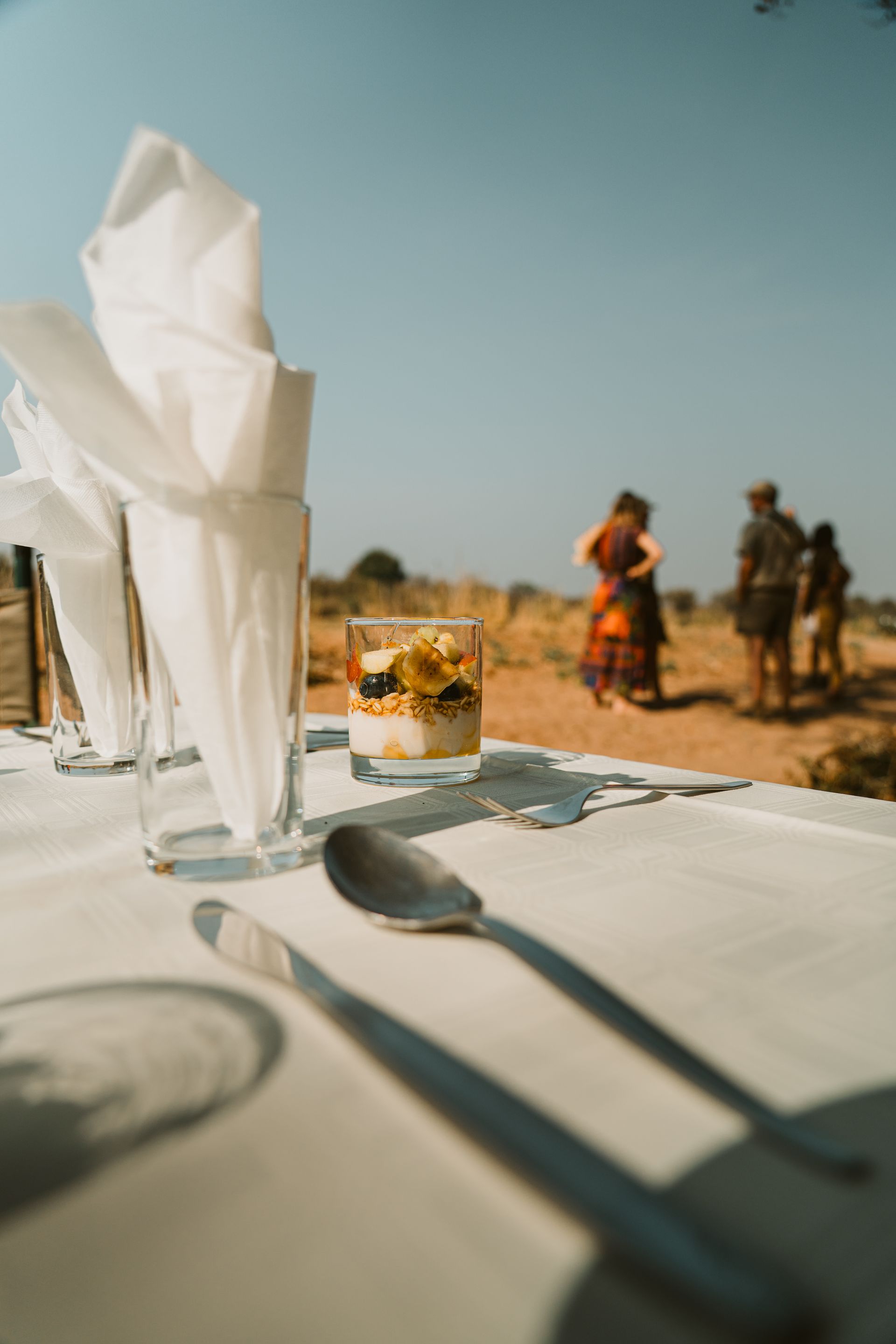 Table set for dining outdoors; glass of parfait, napkins, silverware, blurred figures in the background.