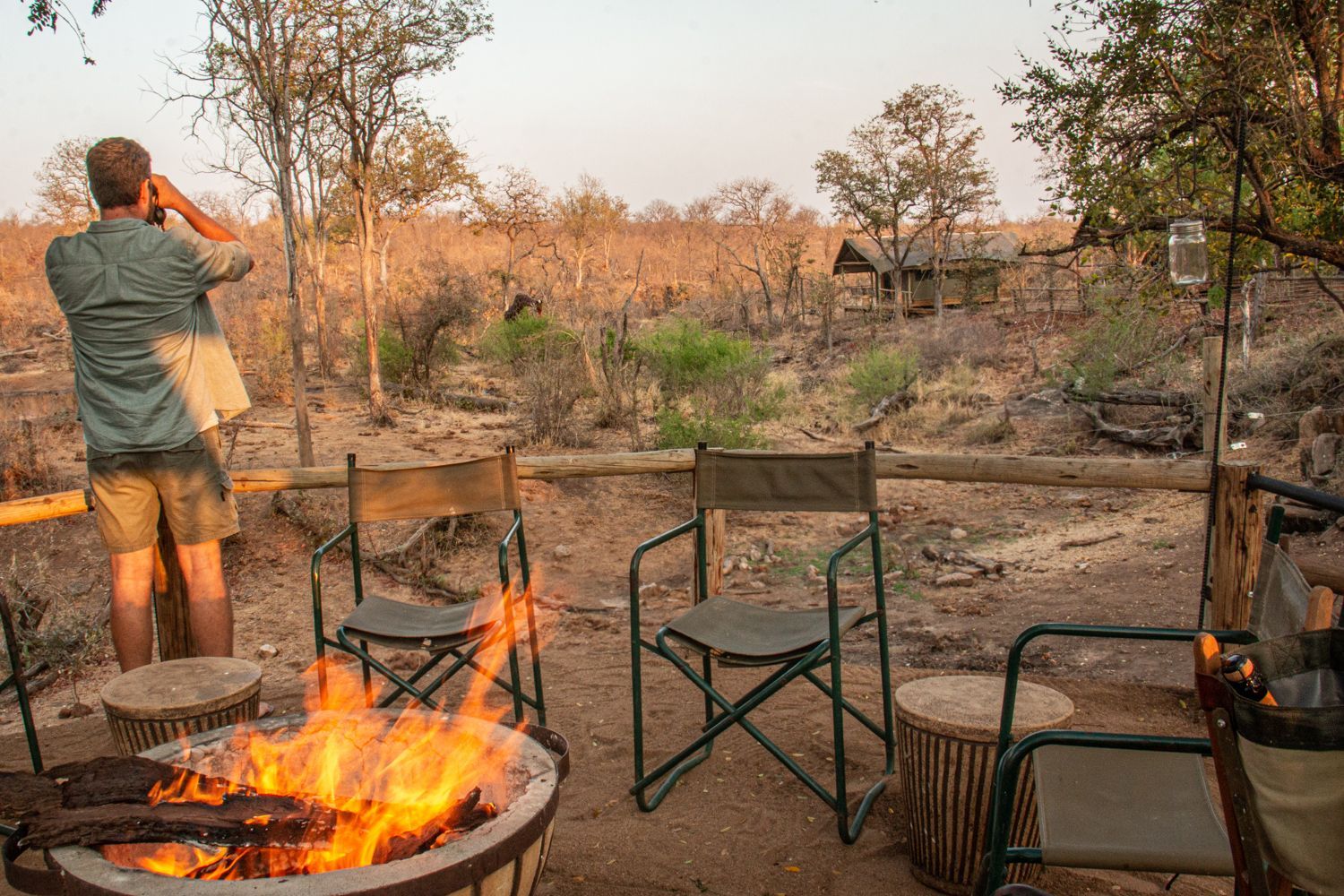 Bundox Safari guest looks through binoculars at a Giraffe near a campfire, chairs. Dry savanna background.