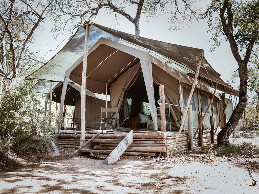 Safari tent on a wooden platform, beige canvas and silver roof, in a wooded area.