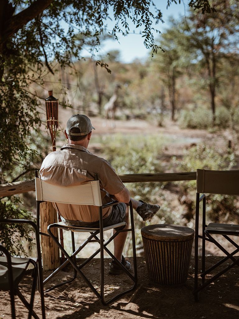 A person in a hat sits in a camp chair outdoors, looking toward a giraffe standing in the brush in the distance.