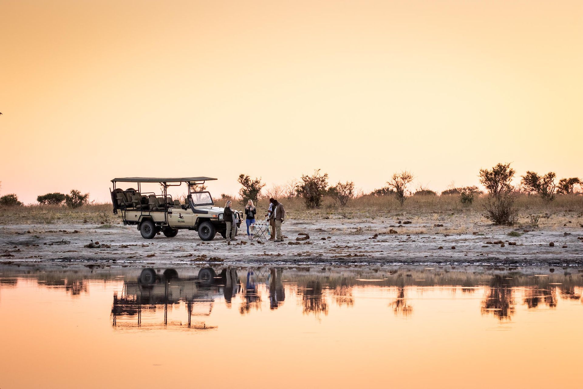 Safari vehicle and people silhouetted at sunset beside a reflective waterhole