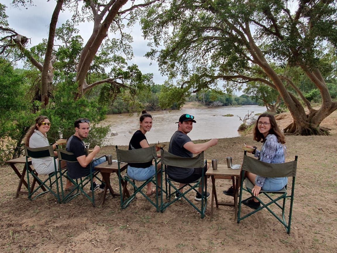 Five people sit in camping chairs by a river under trees, smiling and chatting.