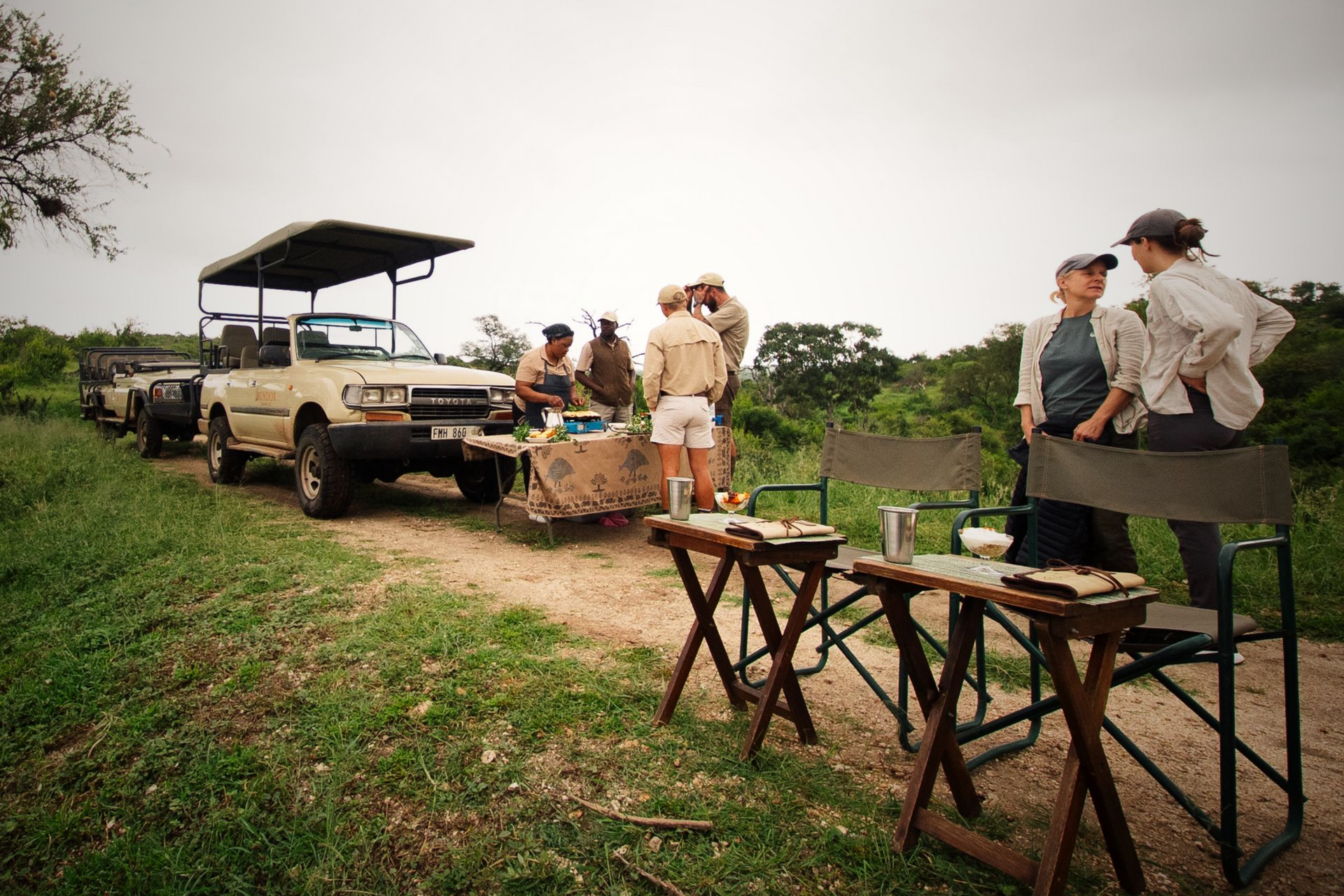 Safari camp with parked trucks, people gathered around folding tables on a grassy plain.