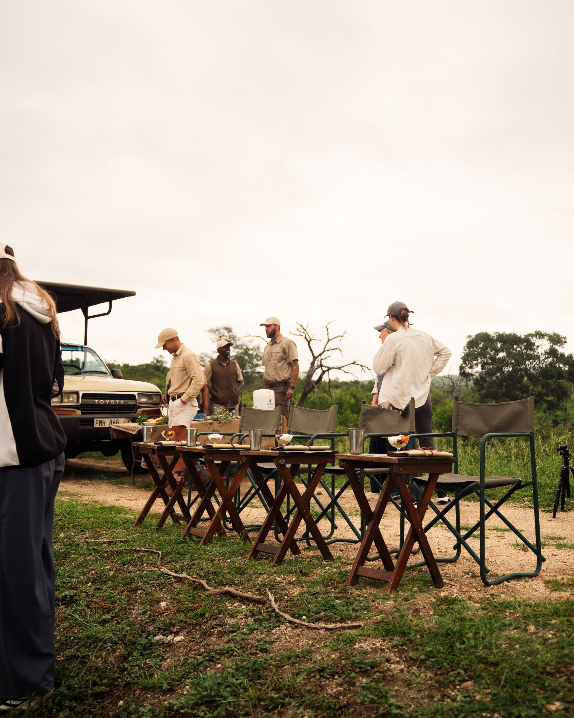 People gathered outdoors near tables in a vineyard, with a vehicle and trees in the background.