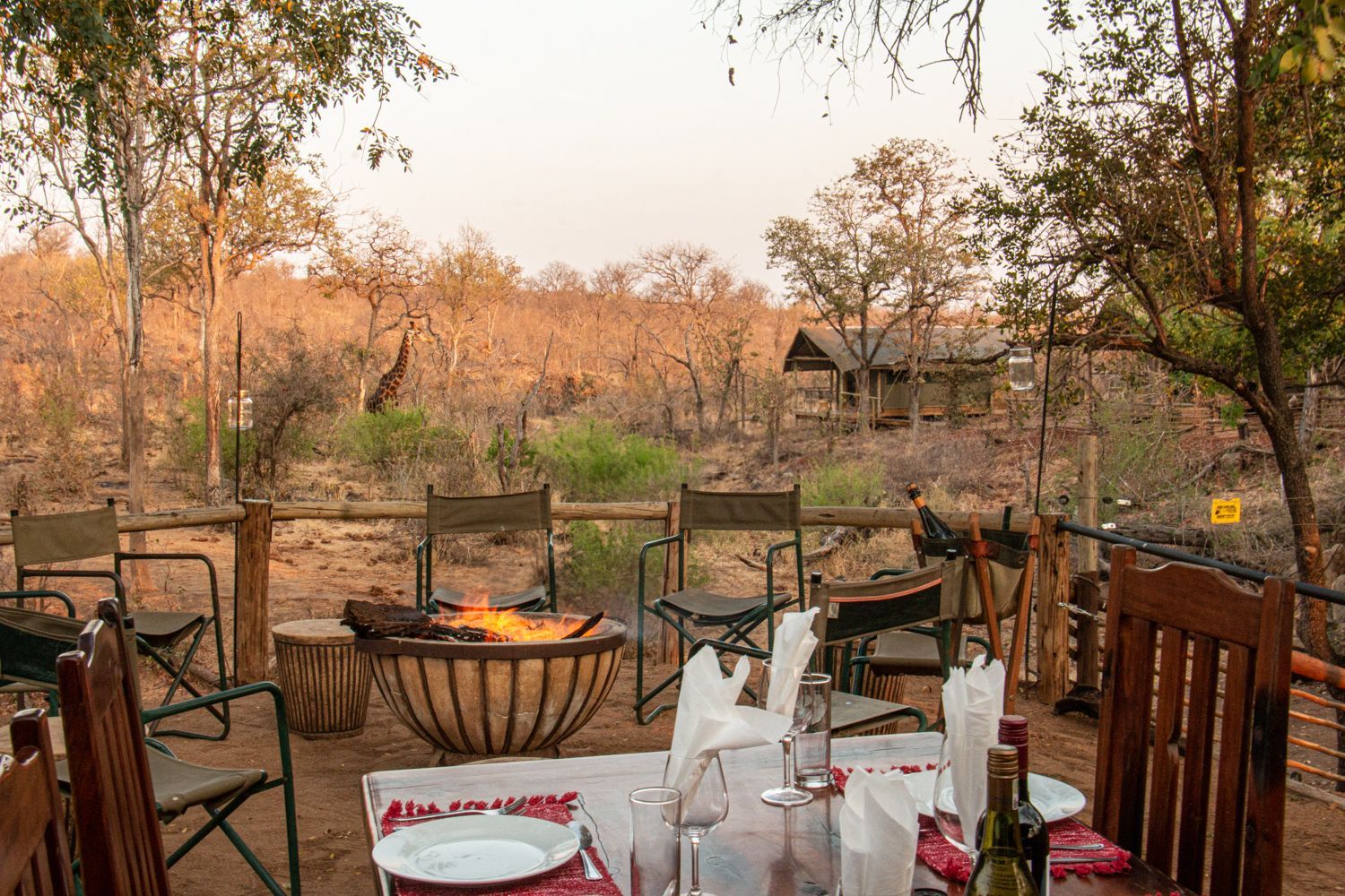 Outdoor dining area with a fire pit and tables overlooking a dry, wooded landscape at sunset