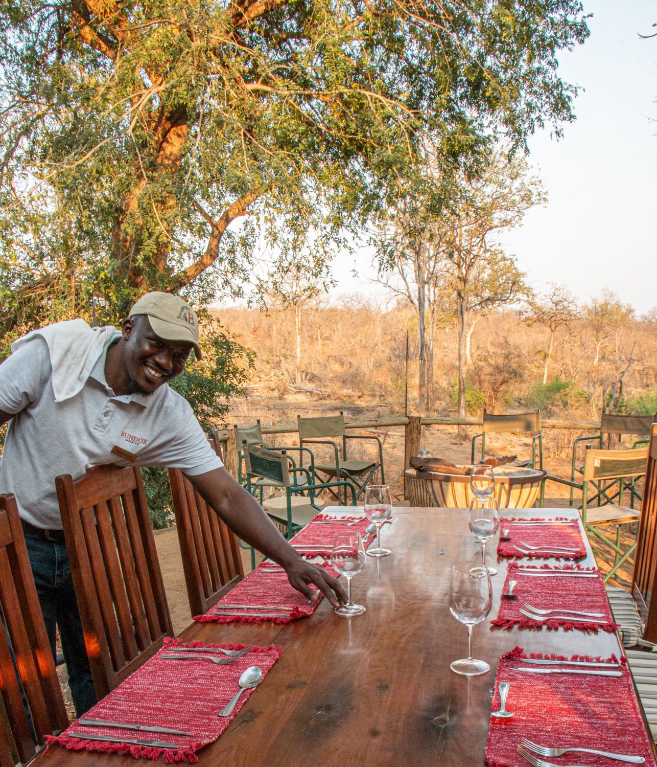 A smiling person wearing a cap sets a table with red placemats and wine glasses in an outdoor safari dining area.