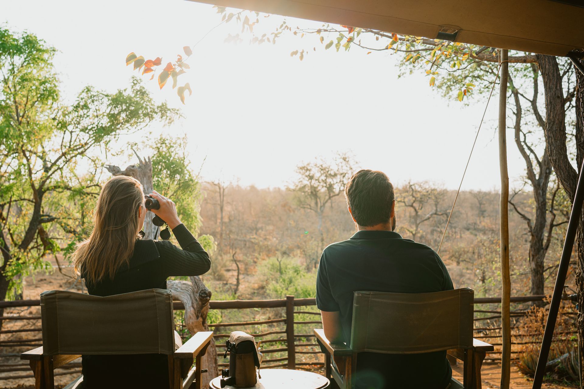 Two people sit in chairs on a shaded wooden deck, looking out over a sunlit savanna landscape with binoculars.