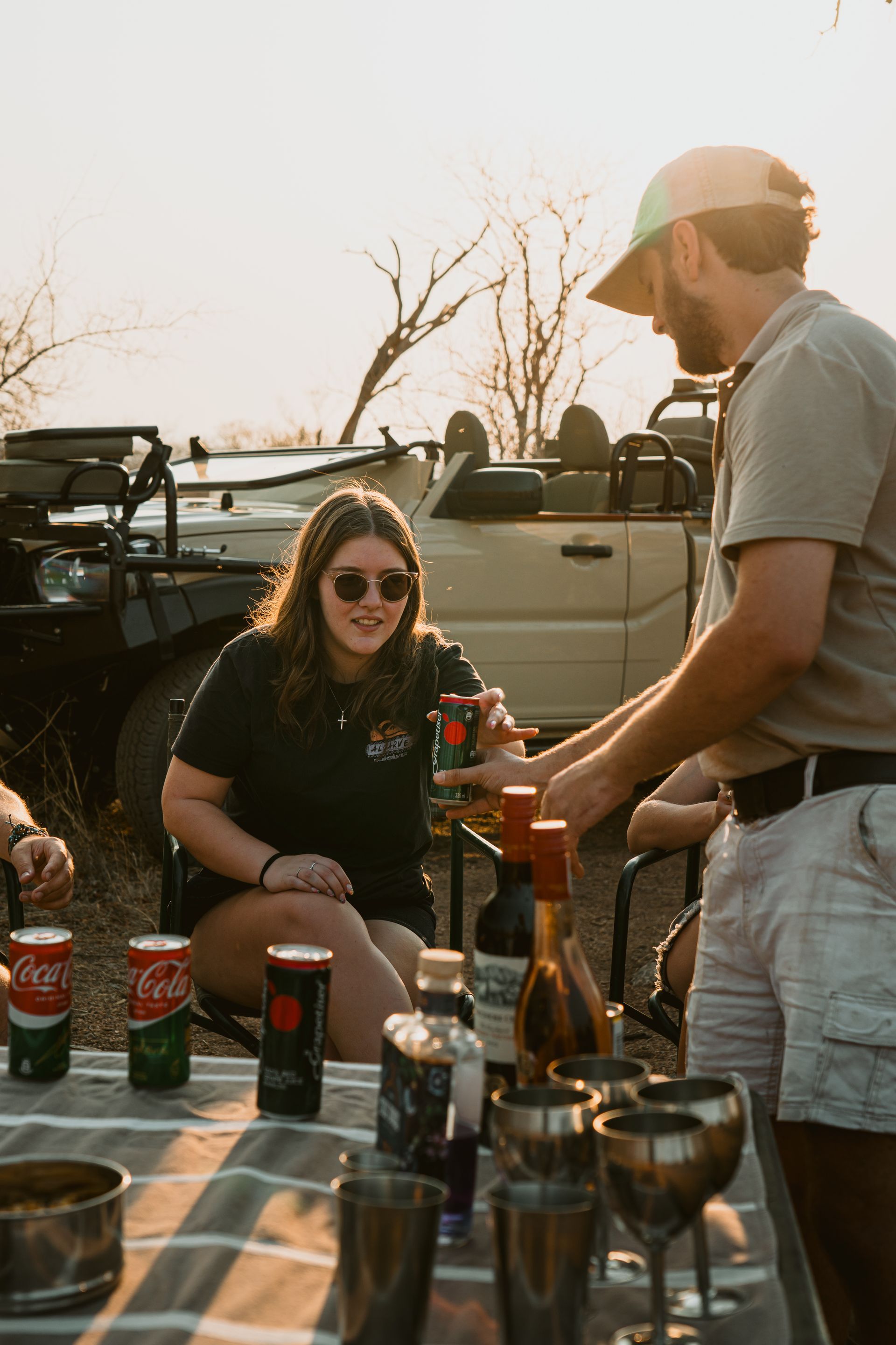 People chatting and grilling outdoors at a picnic table with drinks and sunglasses at sunset