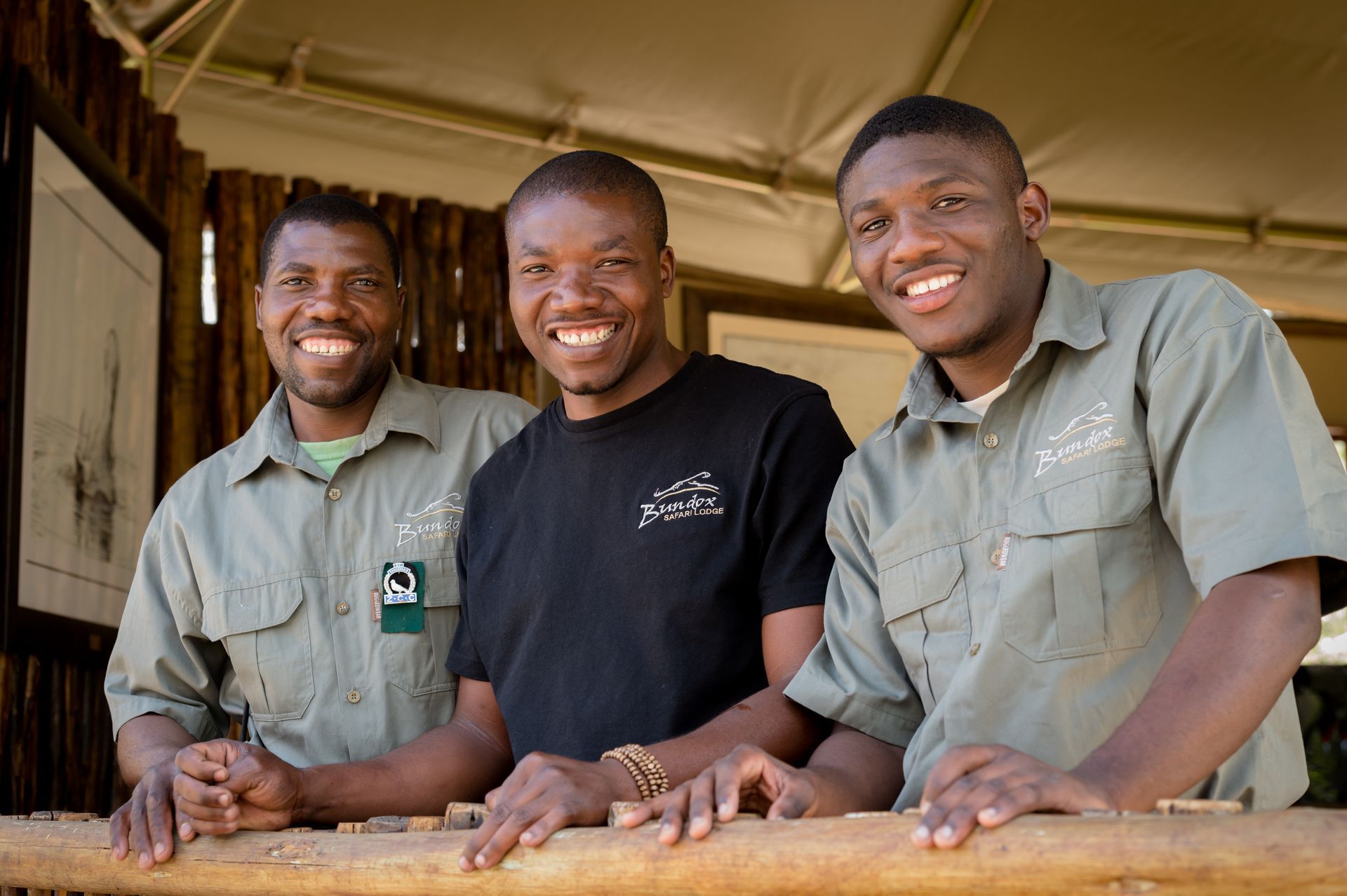 Three smiling workers in green uniforms leaning on a wooden rail outdoors