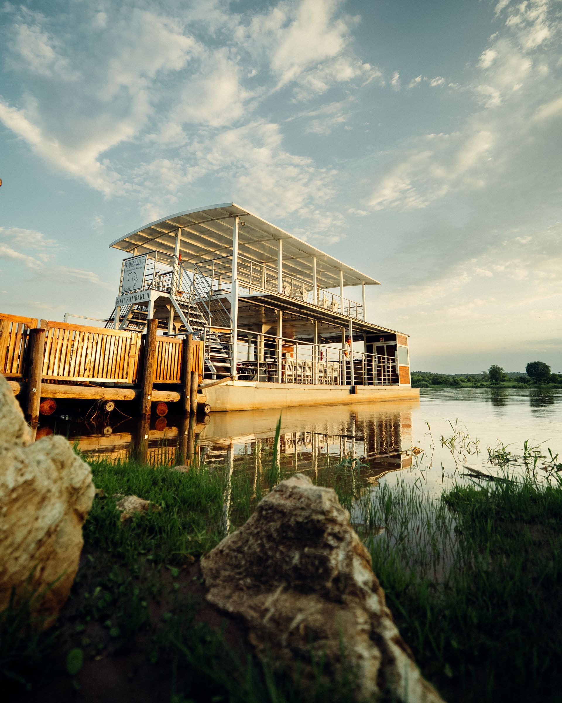 Rustic riverside pavilion at sunset, with wooden structure reflected in calm water and grassy foreground