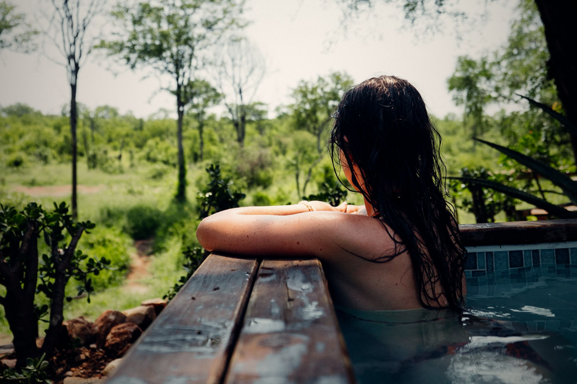 Person relaxing in a pool, looking over a wooden railing toward a lush green landscape.