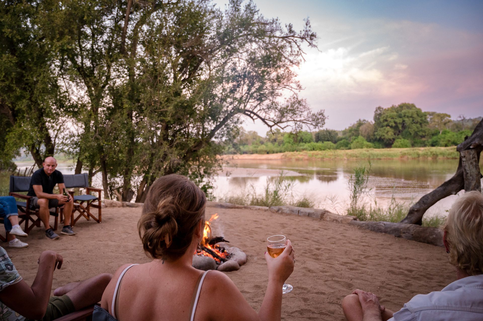 People relaxing by a campfire beside a calm river at sunset, with trees and chairs around them