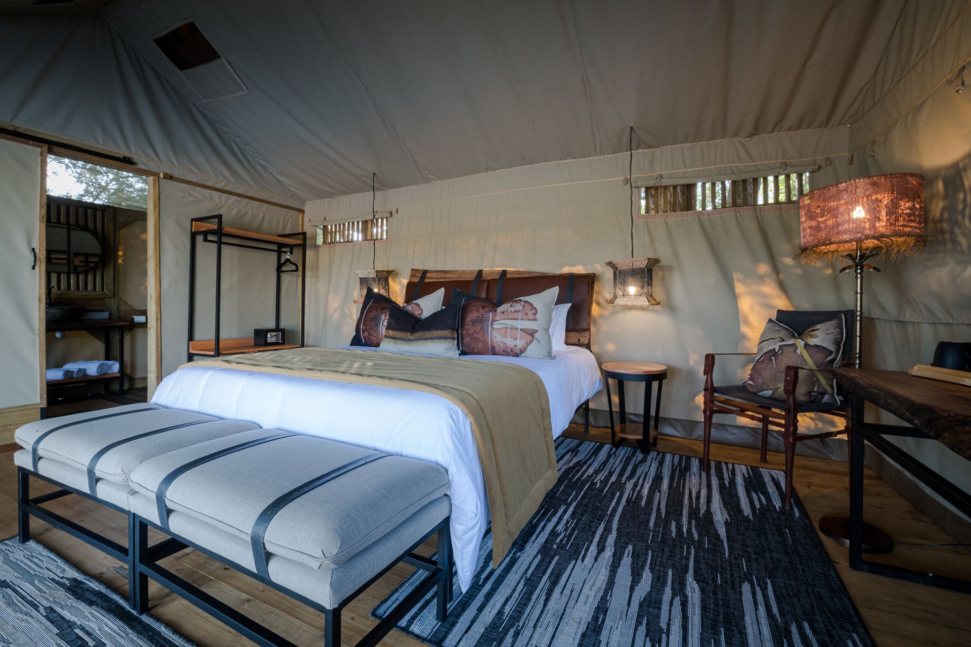 Safari-style bedroom with a king bed, patterned bench, and warm lamp-lit decor under a tented ceiling