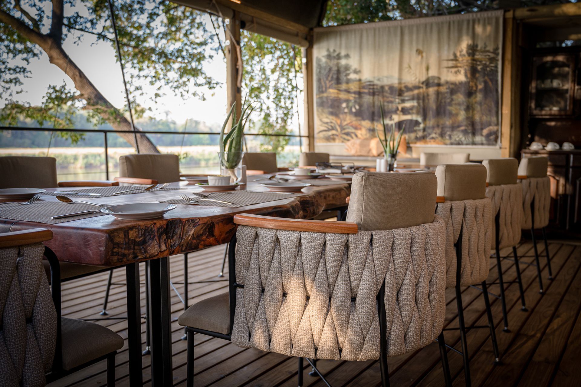 Open-air dining terrace with set tables, woven chairs, and a scenic wall mural.