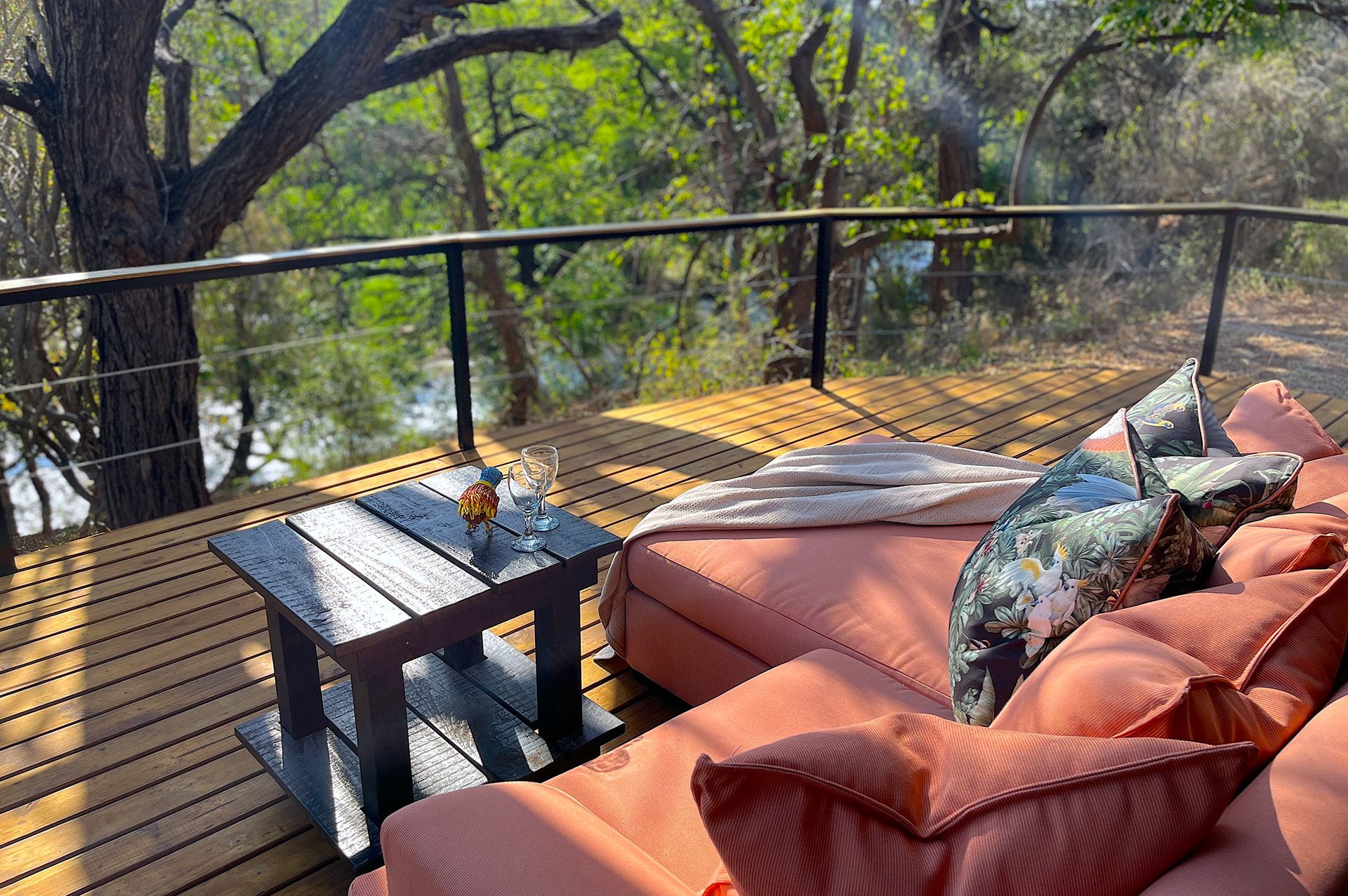 Outdoor deck with salmon-colored seating, black table, and glass railing overlooking a river and trees.