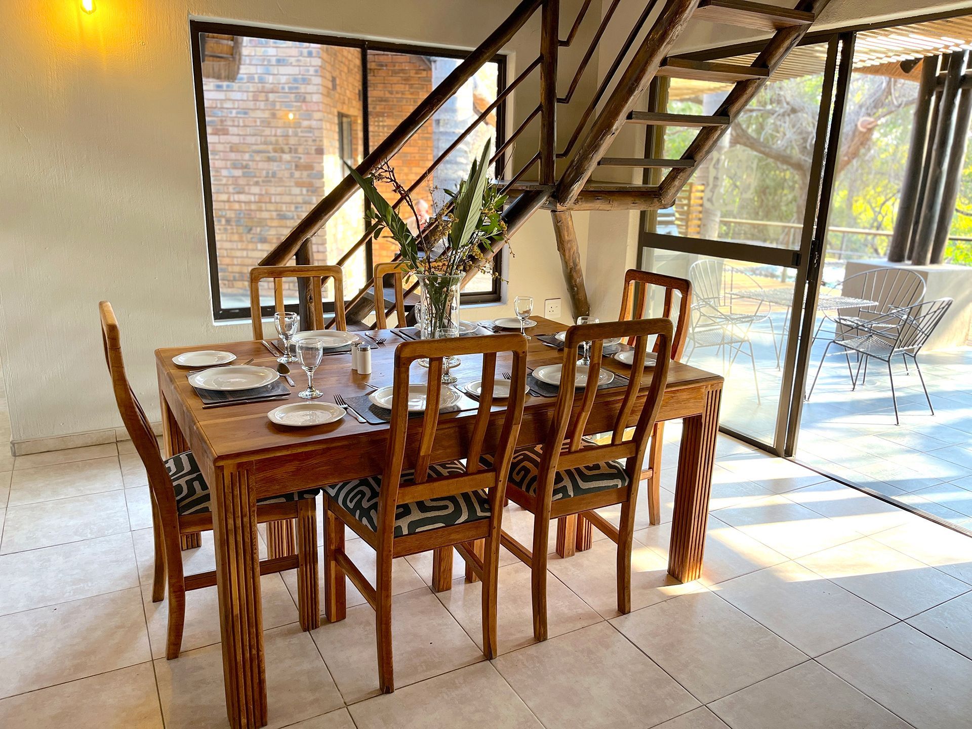 Dining room with wooden table and chairs set for a meal, with stairs and large windows.
