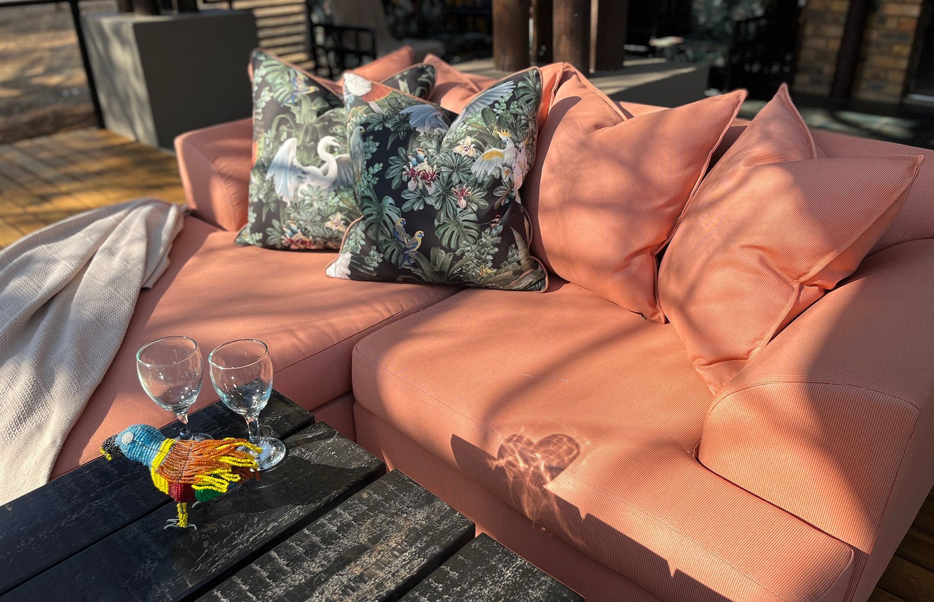 Outdoor patio sofa with coral cushions, patterned pillows, and a black table with drinks in sunlight