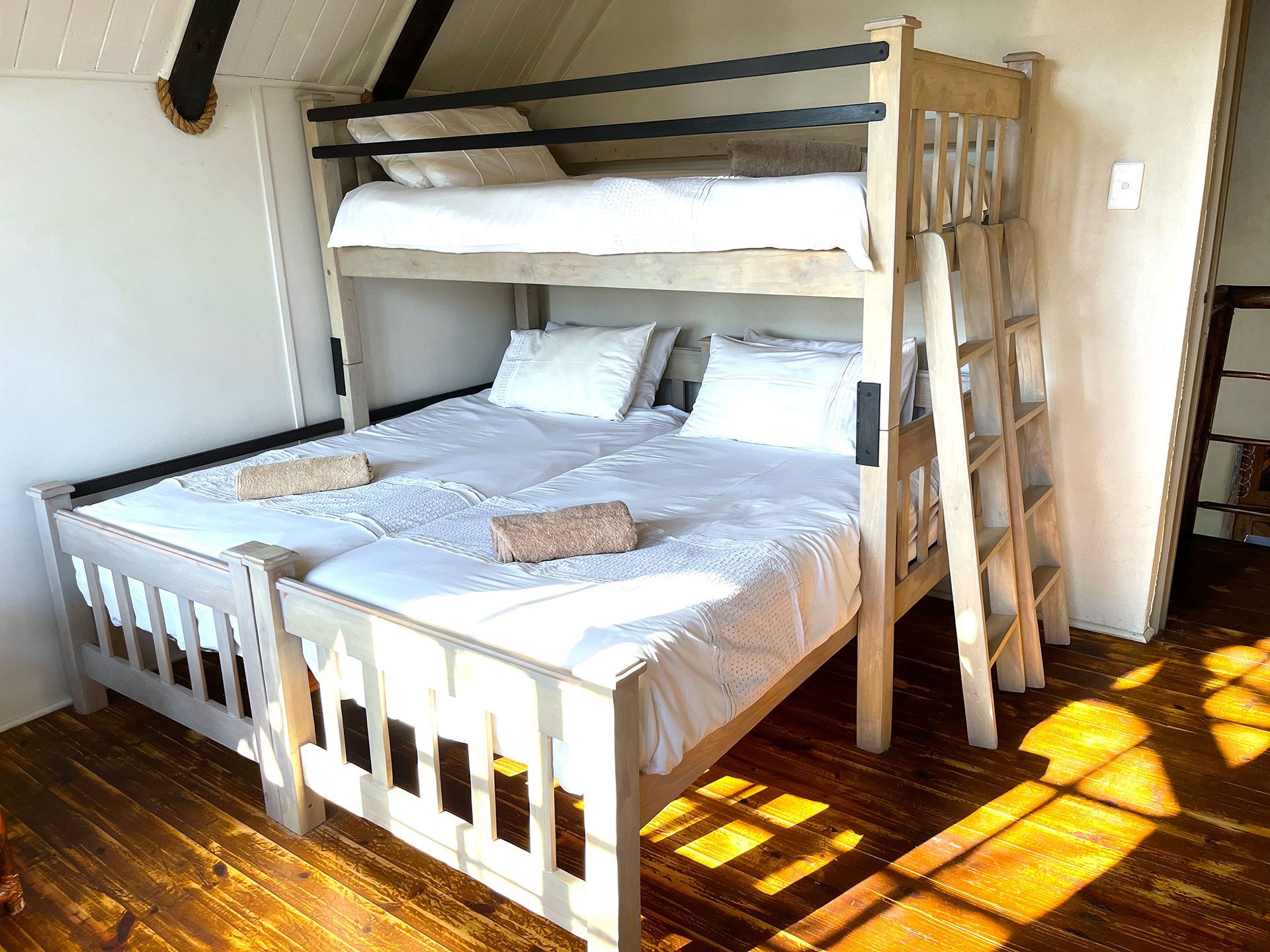 Bunk bed in a room. Lower bed is two twins, top is a twin. Wooden frame, white bedding. Sunlight streams in.