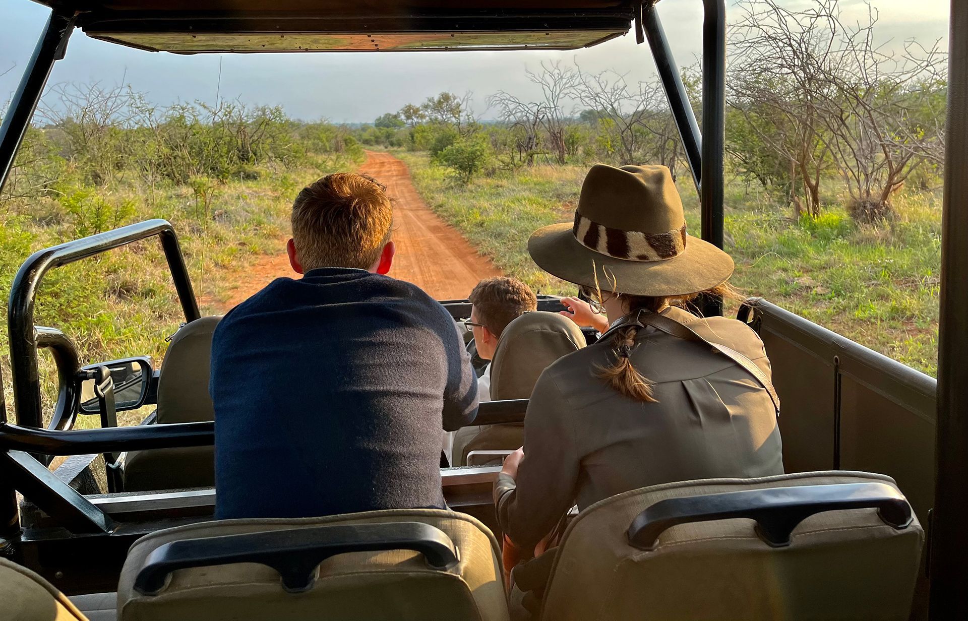 Two people ride in an open safari vehicle on a dirt path through dry grassland.