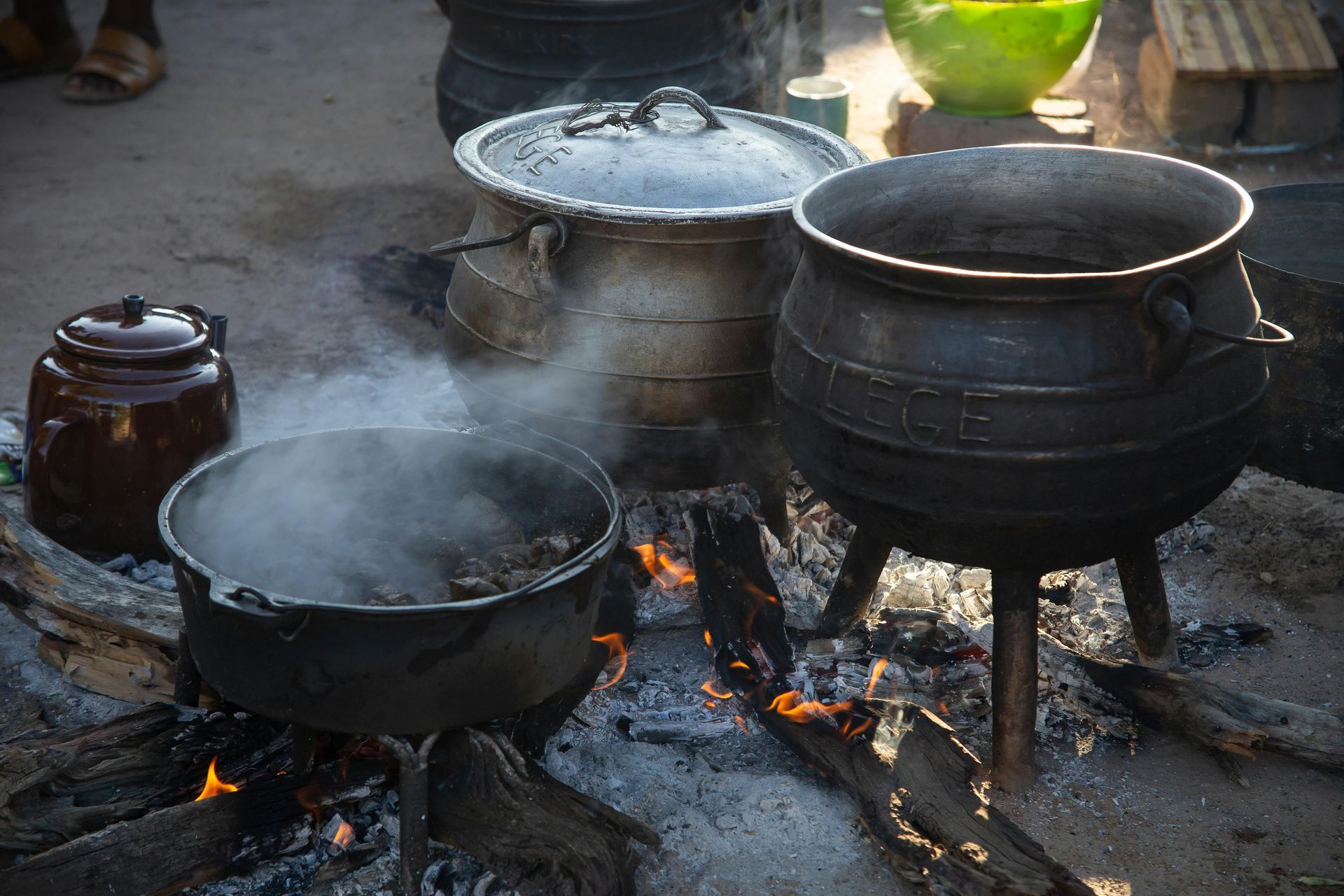 Pots cooking over a fire, outdoors. Smoke rises from the food in the cast iron pots.